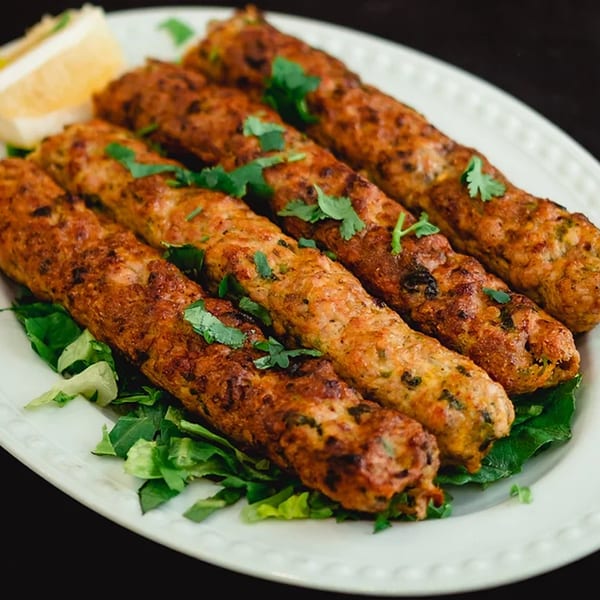 Five Zaffron Afghan seekh kebabs, garnished with cilantro and a lemon wedge, on a white plate.